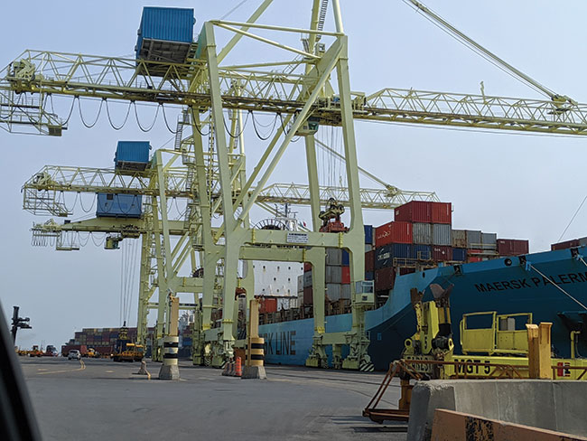 Photograph of a cargo ship loaded with cargo containers that is docked at a marine port, along with large cranes to offload the containers